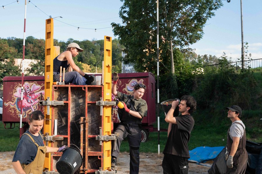 Skupina ľudí dusá hlinu do vysokej debnenej formy počas workshopu